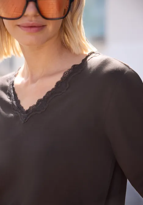 Vrouw met blond bobkapsel en zonnebril draagt een donker T-shirt met V-hals en kanten rand