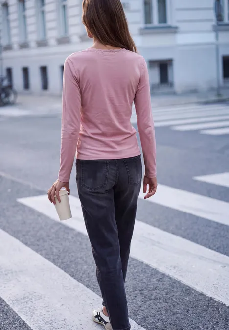 Frau in rosa Oberteil und dunkler Hose quert eine Straße mit Kaffee in der Hand.