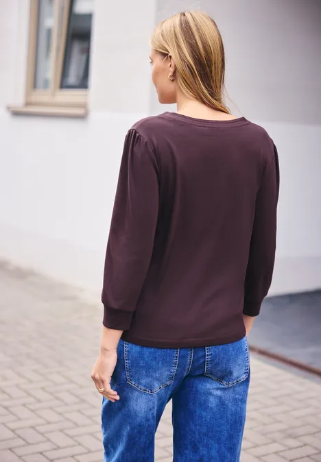 Een vrouw van achteren, gekleed in een donkere top en blauwe jeans, staat in een stedelijke omgeving.