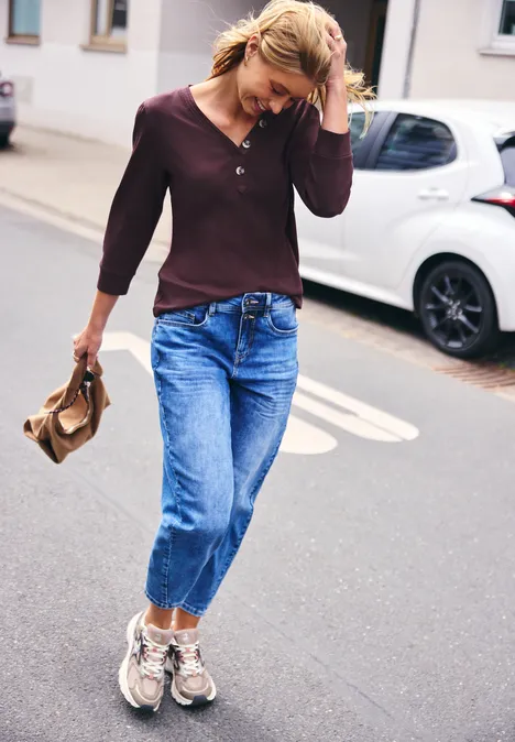 Jonge vrouw loopt over straat, draagt jeans, donker shirt, sneakers en houdt een hoed vast.