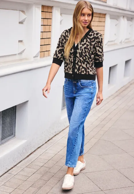 Junge Frau im Leoparden-Cardigan, blauen Jeans und cremefarbenen Loafers auf dem Gehweg.