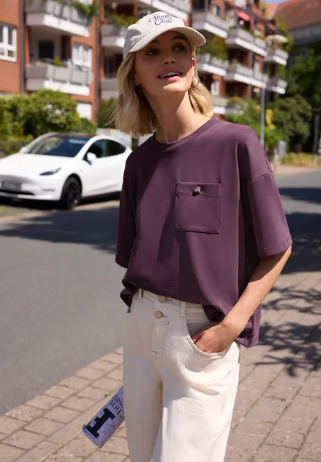 Person in lila T-Shirt und hellen Hosen steht entspannt mit Hand in Tasche auf dem Bürgersteig einer Wohnstraße.