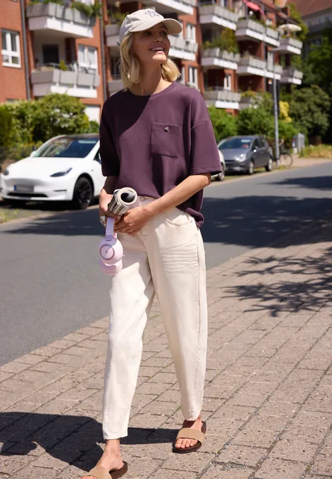 Frau in lila T-Shirt, heller Hose und Cap steht mit Kamera auf dem Gehweg vor Wohnhäusern.