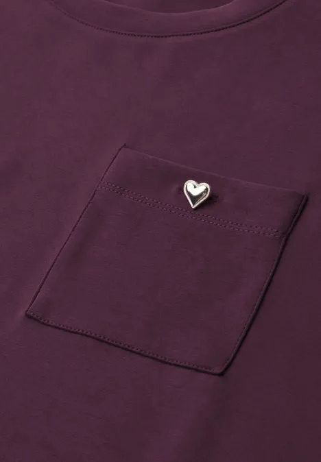 Lila T-Shirt mit Brusttasche und kleinem silbernem Herz-Anstecker.