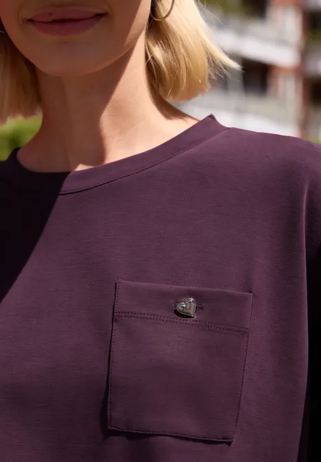 Frau in einem dunkellila Shirt mit aufgesetzter Brusttasche und kleinem goldenen Emblem.