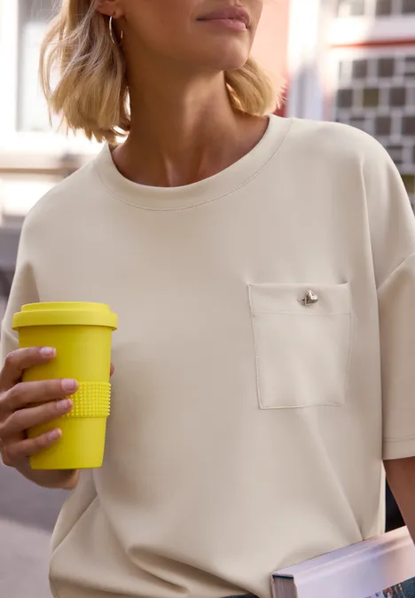T-Shirt im Silk-Look mit Brusttasche creamed sand Eine Person in beigem Oberteil mit Brusttasche hält einen gelben Coffee-to-go-Becher.