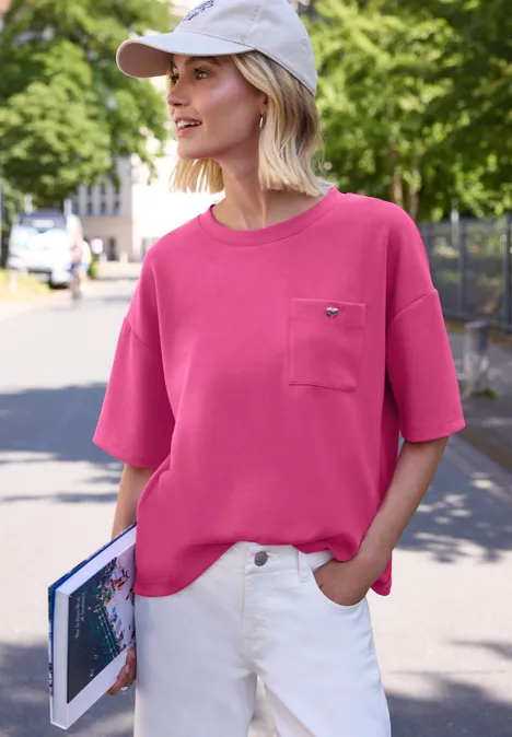Eine Frau in rosa T-Shirt, weißer Hose und Cap hält ein Buch und läuft draußen.
