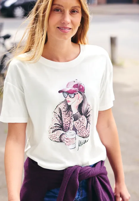 Lächelnde Frau mit weißem T-Shirt und lila Pullover um die Taille, steht draußen und schaut in die Kamera.