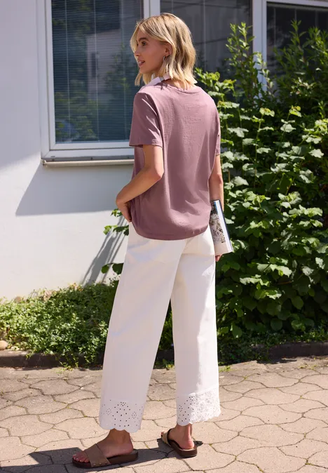 Eine Person in rosa T-Shirt und weißen, weiten Hosen mit Spitzenbesatz steht vor einem Haus und hält eine Tasche.