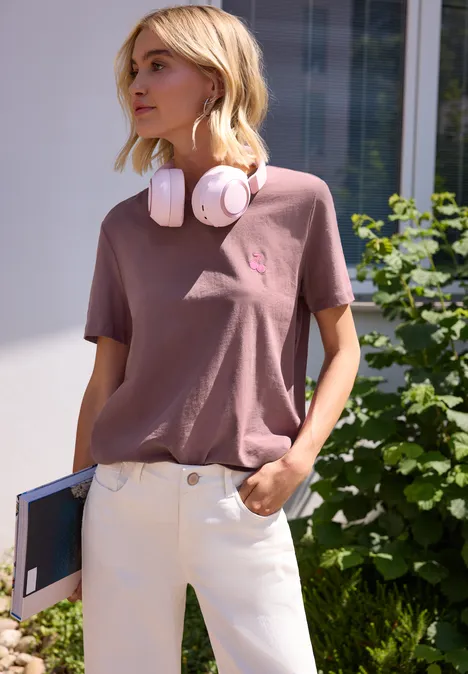 Person mit rosa T-Shirt, weißen Hosen und Kopfhörern hält ein Notizbuch vor grünen Pflanzen.