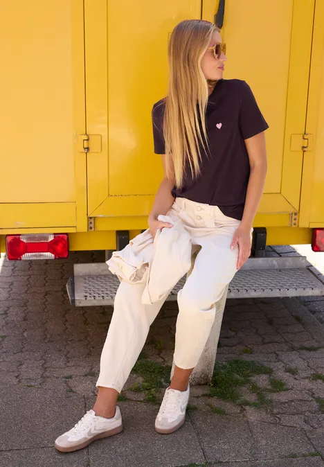 T-Shirt mit Rundhals und Embroidery-Detail very berry Frau lehnt entspannt an gelbem LKW, trägt Sonnenbrille, schwarzes T-Shirt, weiße Hose und Sneaker.
