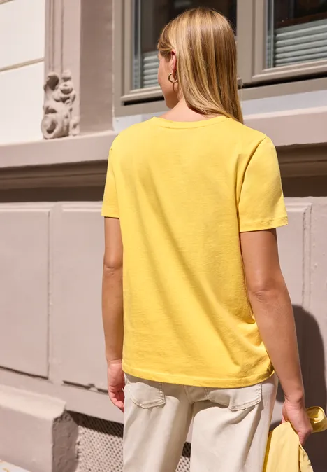 T-Shirt mit Rundhals und Embroidery-Detail snap yellow Eine blonde Frau in gelbem T-Shirt steht mit dem Rücken zur Kamera vor einer Hausfassade.
