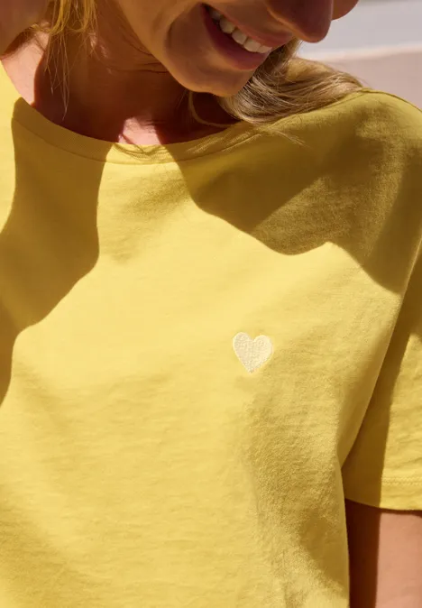 T-Shirt mit Rundhals und Embroidery-Detail snap yellow Nahaufnahme einer Frau im gelben T-Shirt mit kleinem weißen Herz darauf.