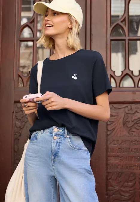 Junge Frau in schwarzem T-Shirt, hellblauer Jeans mit Zierknöpfen und Cap hält Kamera vor einer verzierten Holztür.