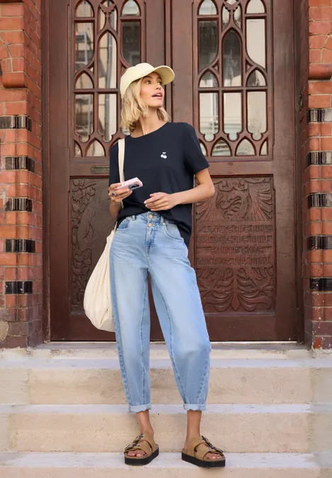 Junge Frau in Jeans, T-Shirt und Hut steht vor einem verzierten Holztor mit Tasche und Handy.