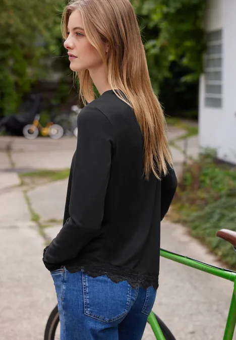 Seitlich blickende Frau mit langem Haar, schwarzem Pullover und Jeans steht draußen neben Fahrrad.