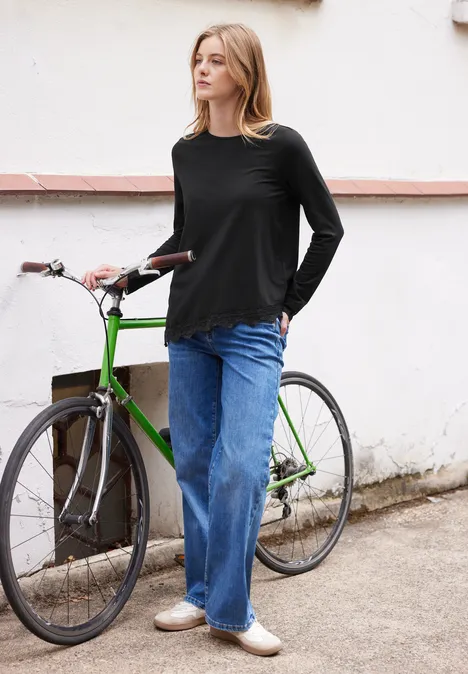 Frau in schwarzem Shirt und blauen Jeans steht neben grünem Fahrrad an weißer Wand.