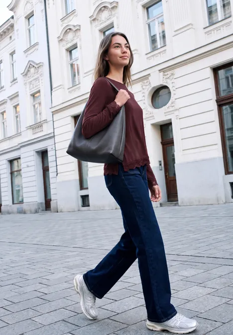 Jonge vrouw loopt in jeans, trui en sneakers met een tas op een stadsstraat.