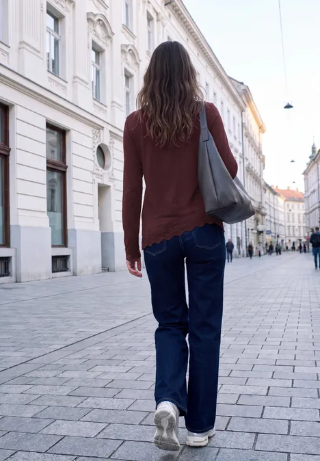 Een vrouw in jeans en bruin shirt met grijze tas loopt over een geplaveide straat.