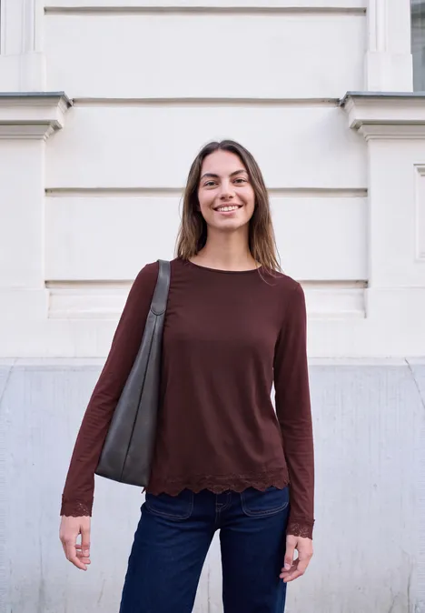 Jonge vrouw in jeans en bordeaux trui, met schoudertas, staand voor een lichte gevel.
