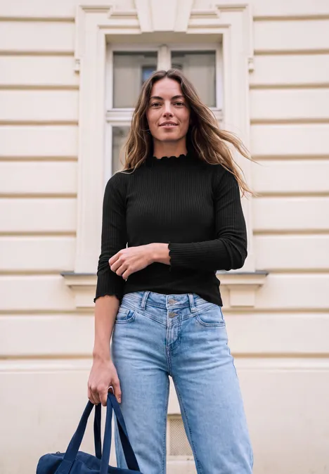 Shirt met lange mouwen en col Black Eine junge Frau steht vor einem hellen Gebäude, trägt schwarzen Pullover, blaue Jeans und hält eine Tasche.