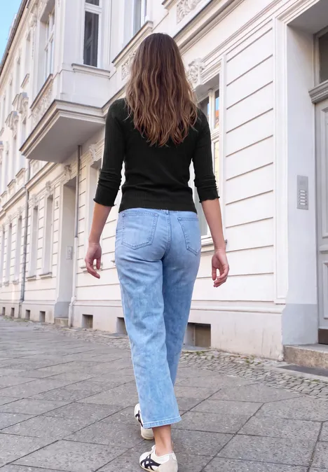 Shirt met lange mouwen en col Black Eine Frau in Jeans und schwarzem Oberteil geht eine Straße mit weißen Häusern entlang.