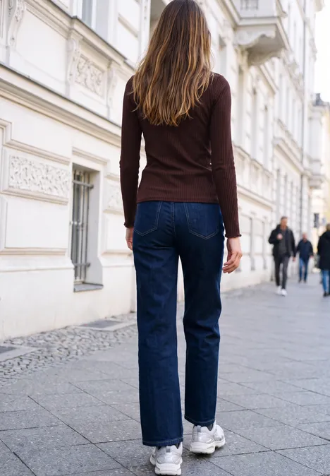 Langarm Shirt mit Turtle Neck russet red Eine Frau in Jeans und Turnschuhen geht eine Pflasterstraße mit historischen Gebäuden entlang.