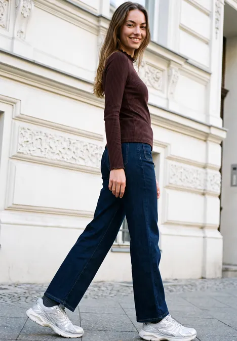 Langarm Shirt mit Turtle Neck russet red Junge Frau in Jeans und braunem Oberteil lächelt beim Spaziergang vor weißer Fassade.