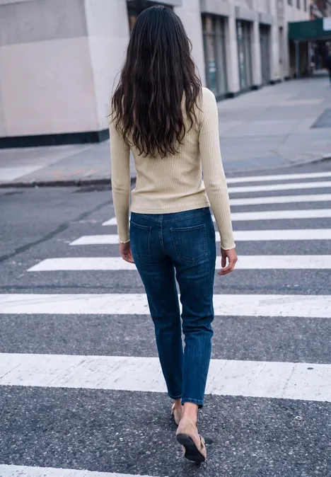 Person mit langen braunen Haaren in Jeans und beigem Pullover überquert Zebrastreifen in der Stadt.