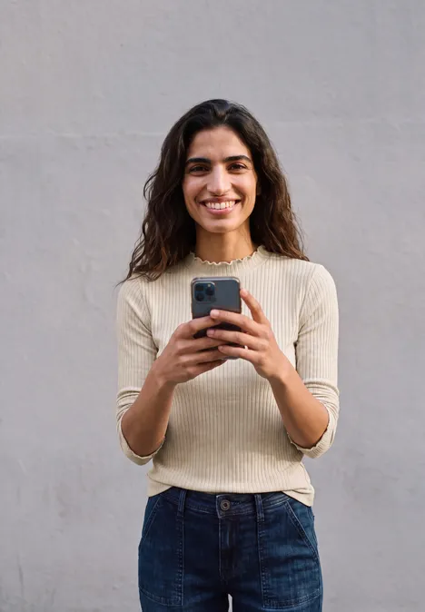 Junge Frau vor einer grauen Wand lächelt und hält ein Smartphone in den Händen.