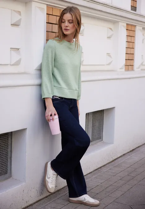 Eine Frau lehnt an einer Wand, trägt hellgrünen Pullover, dunkle Jeans, weiße Sneaker und hält einen Becher.