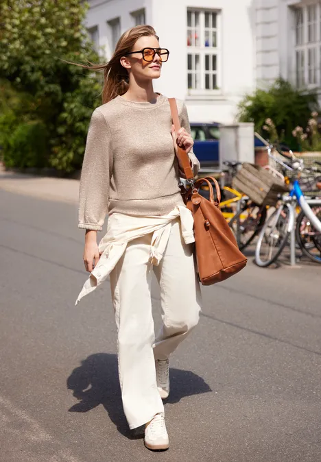 Eine junge Frau in hellem Pullover, weißen Hosen und Sonnenbrille schlendert mit Tasche an sonniger Straße entlang.