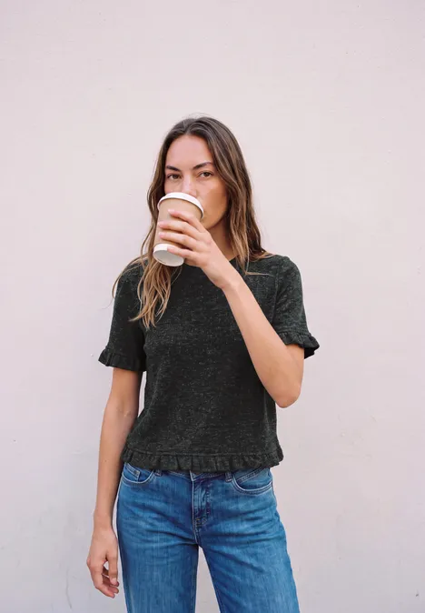 Kurzarm Shirt mit Rundhals und Rüschen inked grey mel. Eine junge Frau trinkt aus einem Pappbecher Kaffee vor einer hellen Wand.