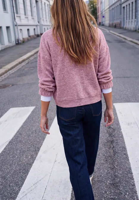 Eine Frau in rosa Pullover und dunklen Jeans überquert von hinten einen Zebrastreifen in der Stadt.