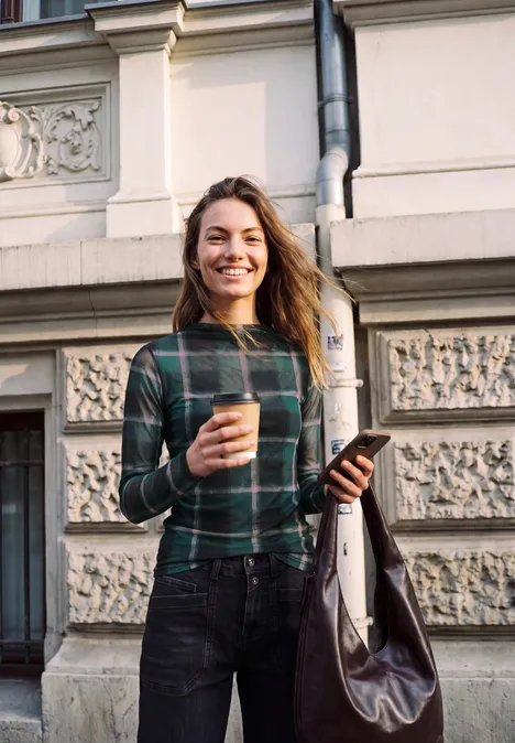 Eine lächelnde Frau im karierten Pullover hält Kaffee und Smartphone vor historischer Hausfassade.