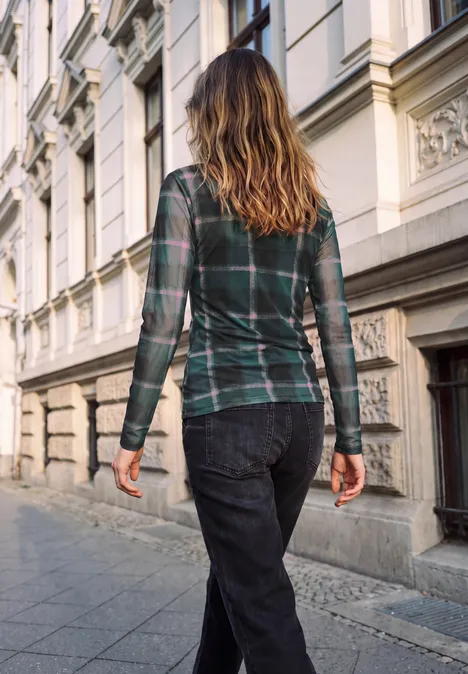 Frau in kariertem Hemd und dunkler Jeans spaziert vor einer Altbau-Fassade davon.