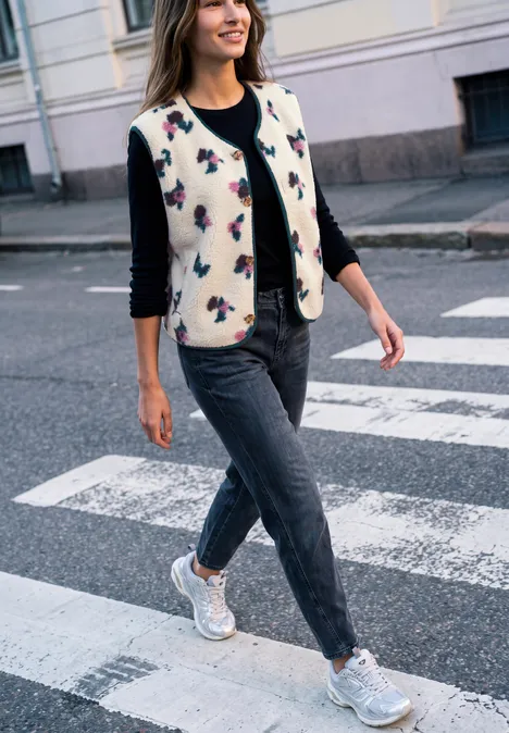 Une femme en jeans, gilet et baskets traverse en souriant un passage piéton en ville.