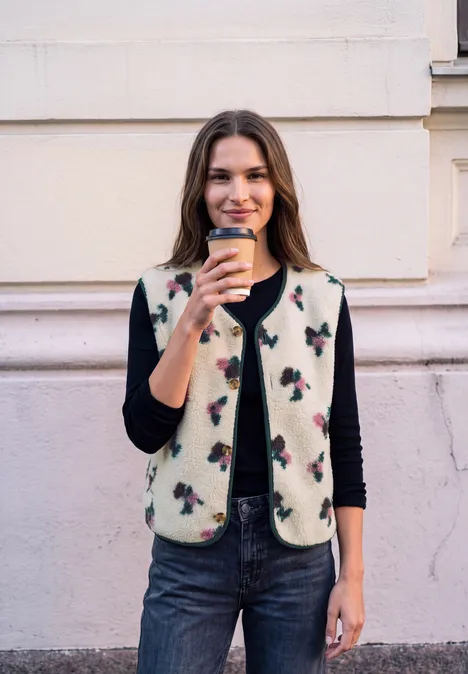 Jeune femme devant un mur, souriante, tenant un café à emporter, vêtue d’une veste à motifs, pull noir et jean.