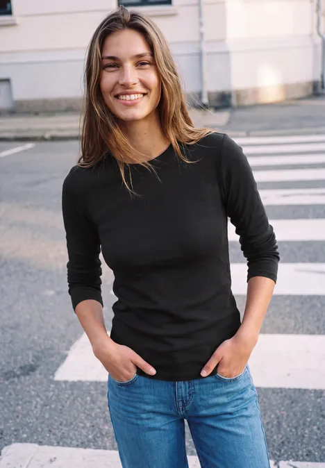 Eine lächelnde junge Frau in schwarzem Oberteil und Jeans steht auf einem Zebrastreifen.