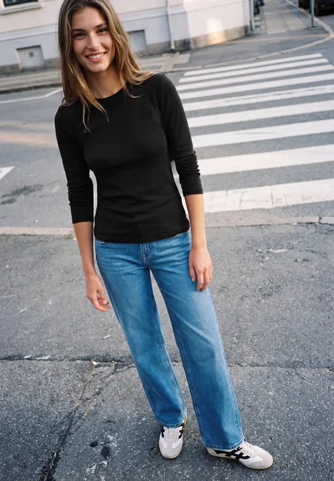 Eine junge Frau in schwarzem Shirt und Jeans lächelt auf einem Zebrastreifen.