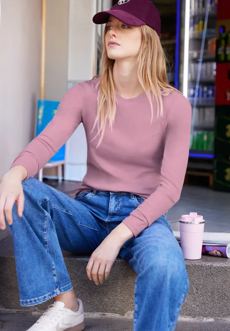 Frau in rosa Shirt und Jeans mit Cap und Trinkbecher sitzt lässig auf einer Treppenstufe.