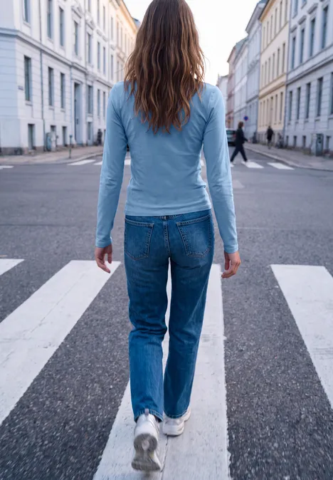 T-shirt basique uni à col rond loom blue Junge Frau in hellblauer Bluse und Jeans überquert einen Zebrastreifen in einer ruhigen Straße mit weißen Häusern.