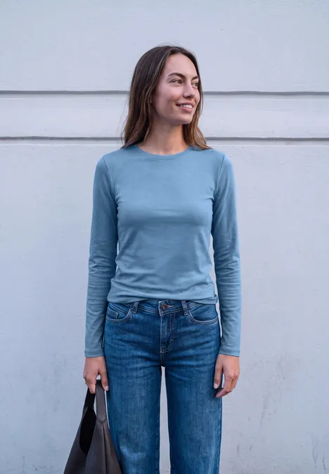 T-shirt basique uni à col rond loom blue Eine lächelnde junge Frau in blauem Shirt und Jeans mit Tasche in der Hand.