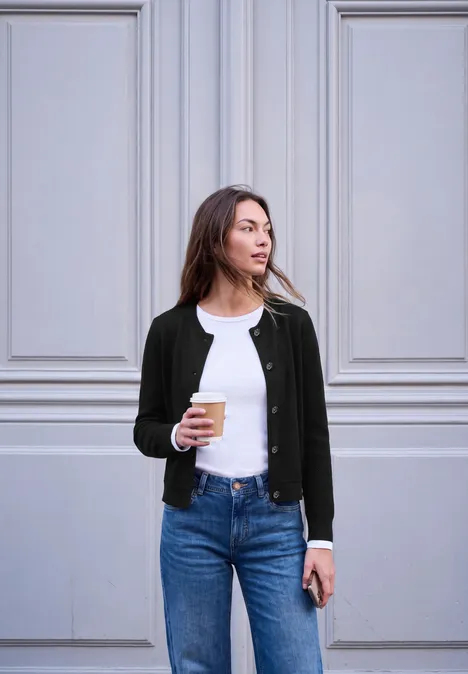 Een vrouw staat voor een lichte muur, draagt een spijkerbroek, t-shirt en vest en houdt koffie en smartphone vast.