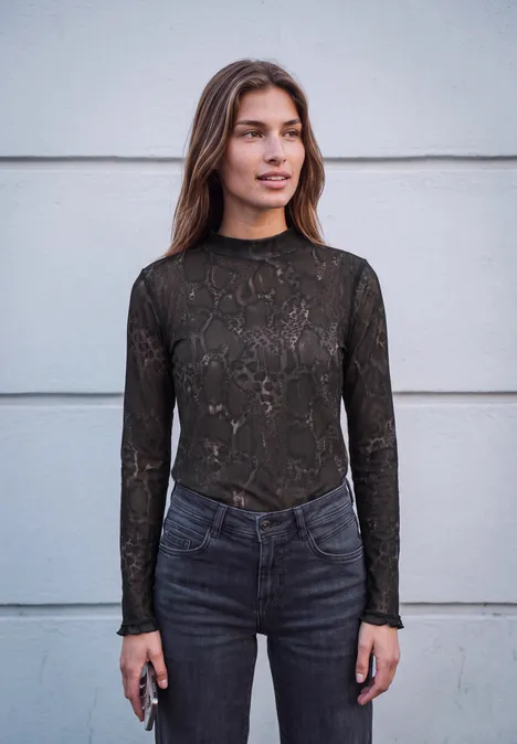 Vrouw met bruin haar voor een grijze muur draagt een donker kanten top en spijkerbroek, houdt een smartphone vast.