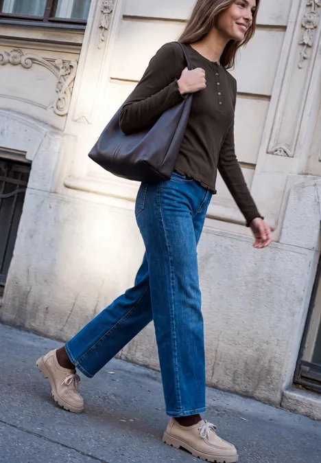 Eine Frau in Jeans, Sneakern und brauner Bluse trägt eine Tasche und läuft an einer Hausfassade vorbei.