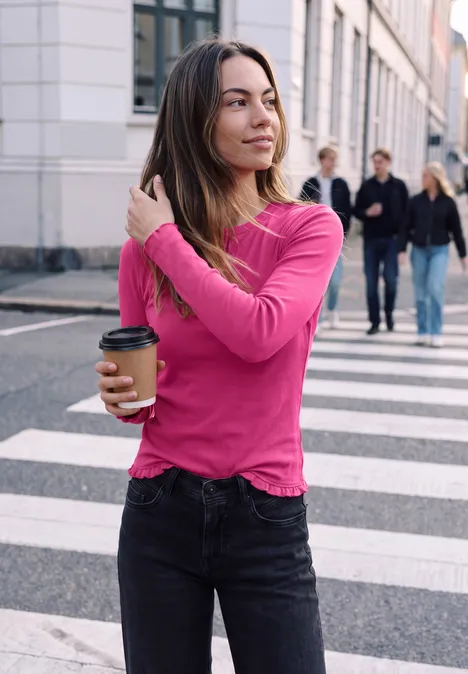 Een vrouw in roze shirt en spijkerbroek loopt met koffie over een zebrapad en strijkt haar haar.