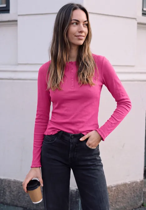 Vrouw in roze lange mouw top en zwarte jeans voor witte muur, met een koffie in haar hand.