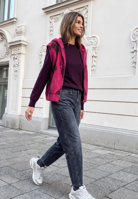 Eine Frau in rosa Jacke und Jeans beim Spaziergang vor einer Gebäudefassade.