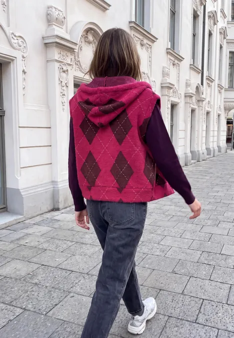 Person geht auf Gehweg vor weißem Haus, trägt pink-gemusterte Kapuzenweste, dunkelgraue Hose und weiße Sneaker